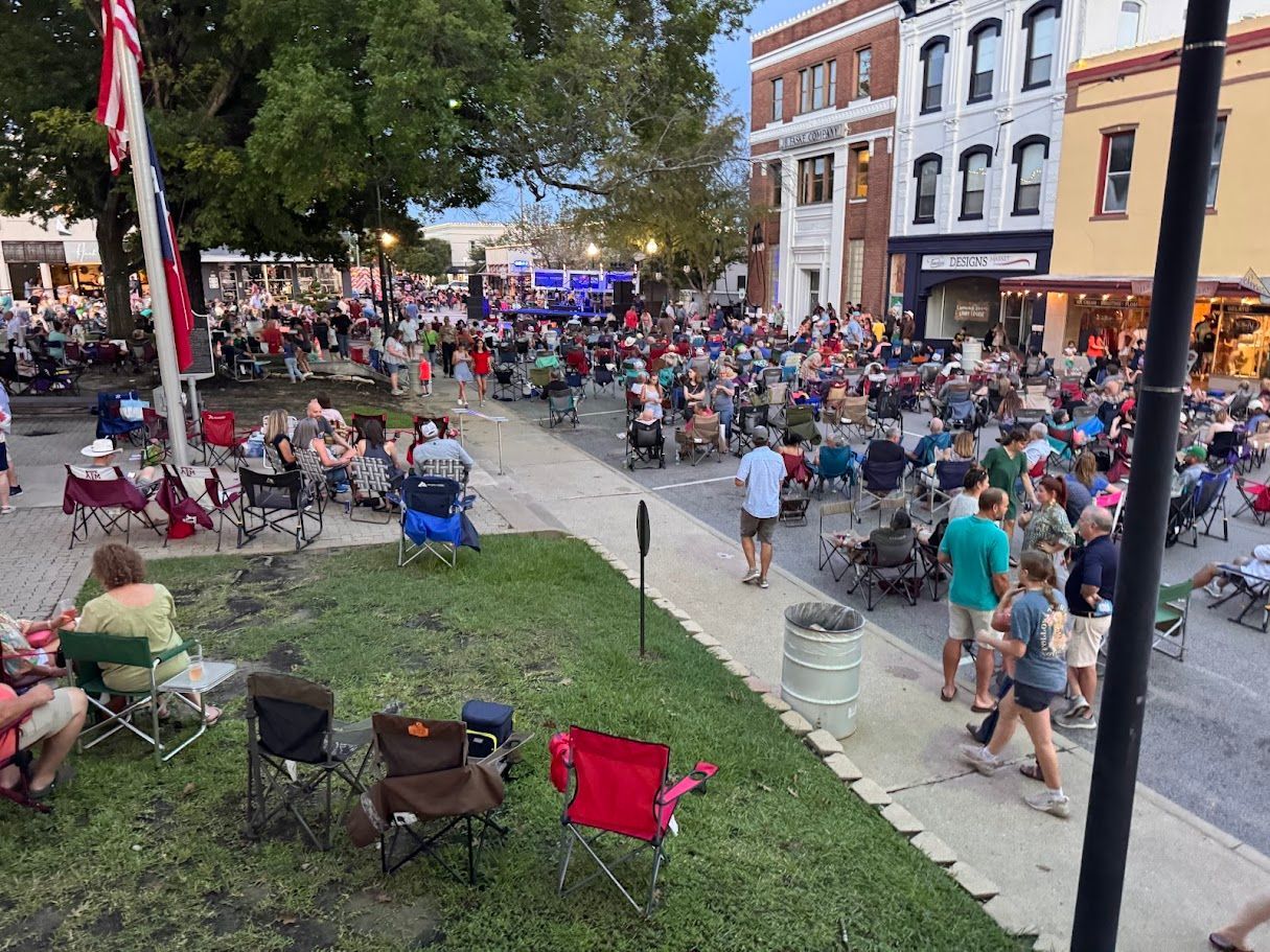 Crowd at outdoor event with stage, buildings, and American flag. People sit in chairs. Evening setting.