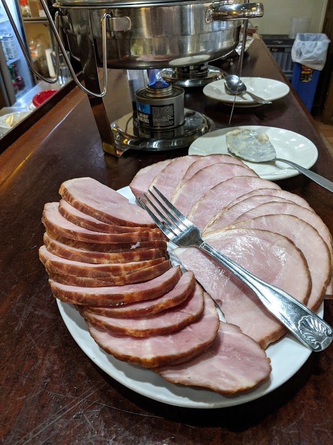 Sliced ham on a white plate with a fork, near a buffet warmer on a wooden counter.
