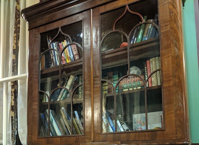Wooden bookcase filled with books behind glass doors, in a room with a window and green wall.