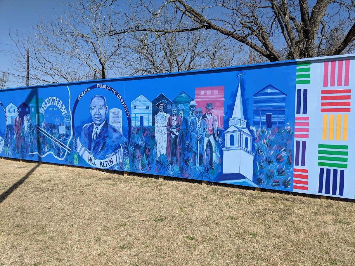 Blue mural with portraits, buildings, and colorful patterns on a grassy area, under a blue sky.