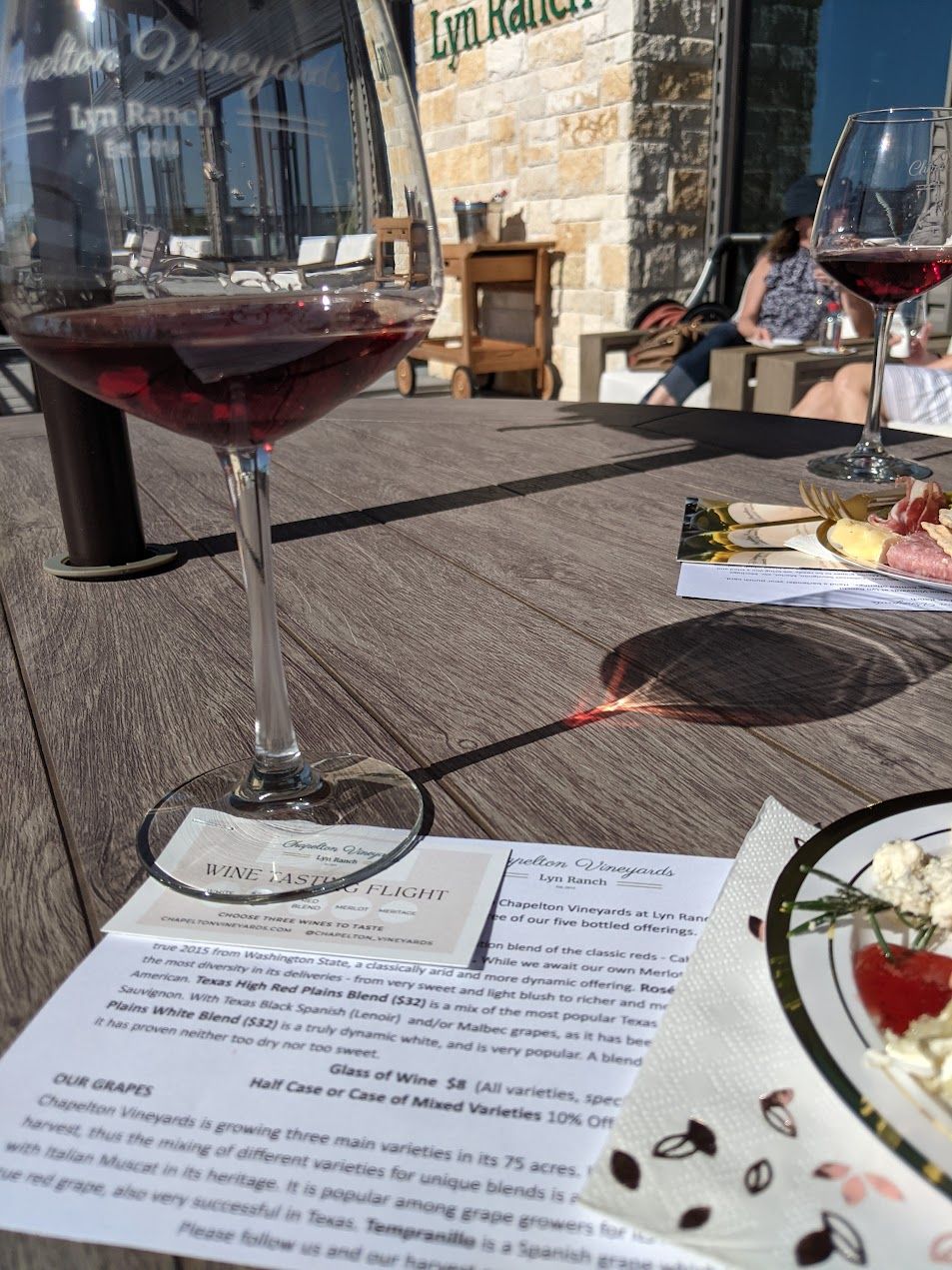 Glass of red wine on a table at a vineyard, with food, papers, and two people in background.