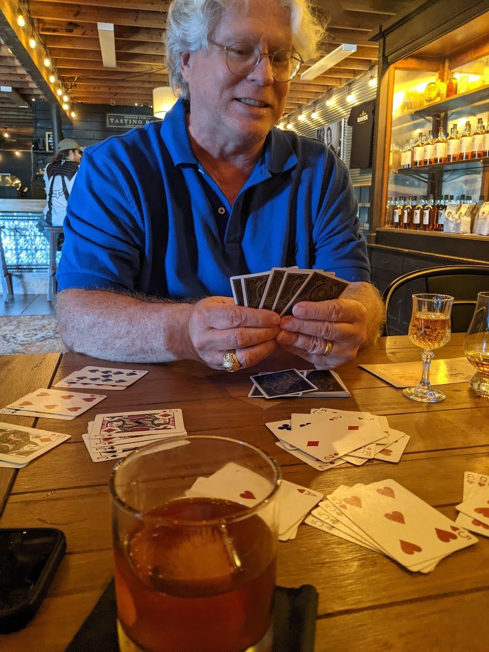 Man playing cards at a wooden table in a bar, holding cards, drink nearby.
