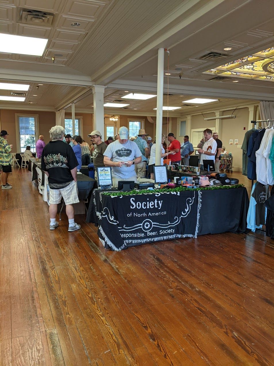 Indoor craft fair with vendors and customers browsing, wooden floor, under white ceiling.