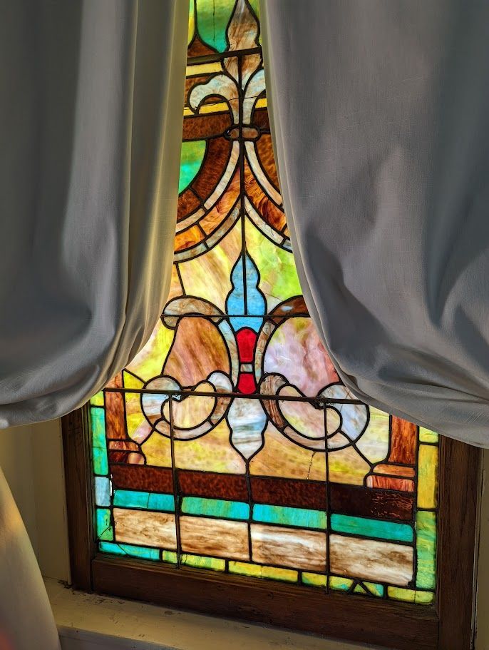Stained-glass window with a fleur-de-lis design framed by white curtains. The colors include blue, red, green, yellow, and brown.