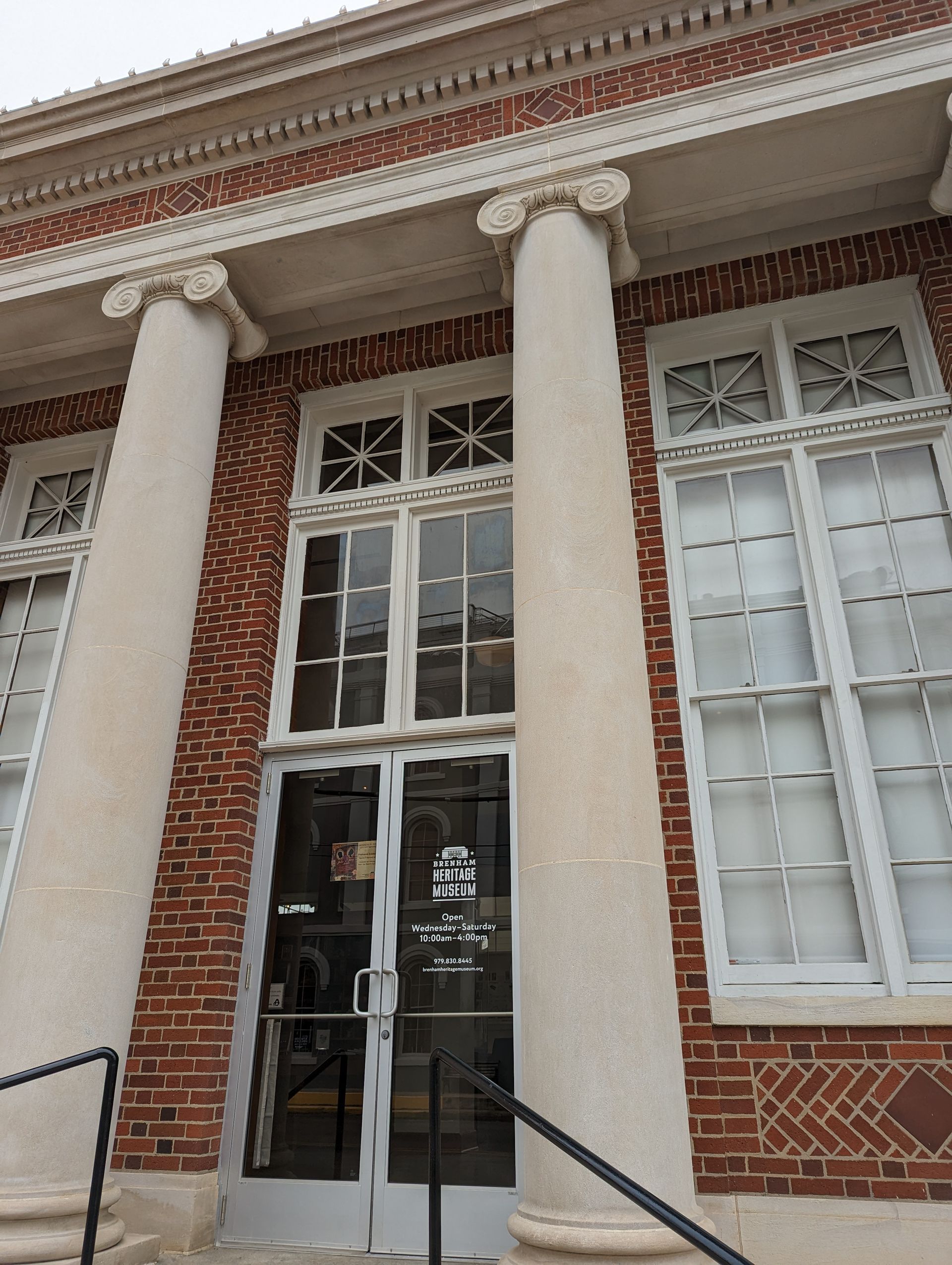 Low-angle shot of a brick building entrance featuring tall white columns with ionic capitals and large multi-paned windows.