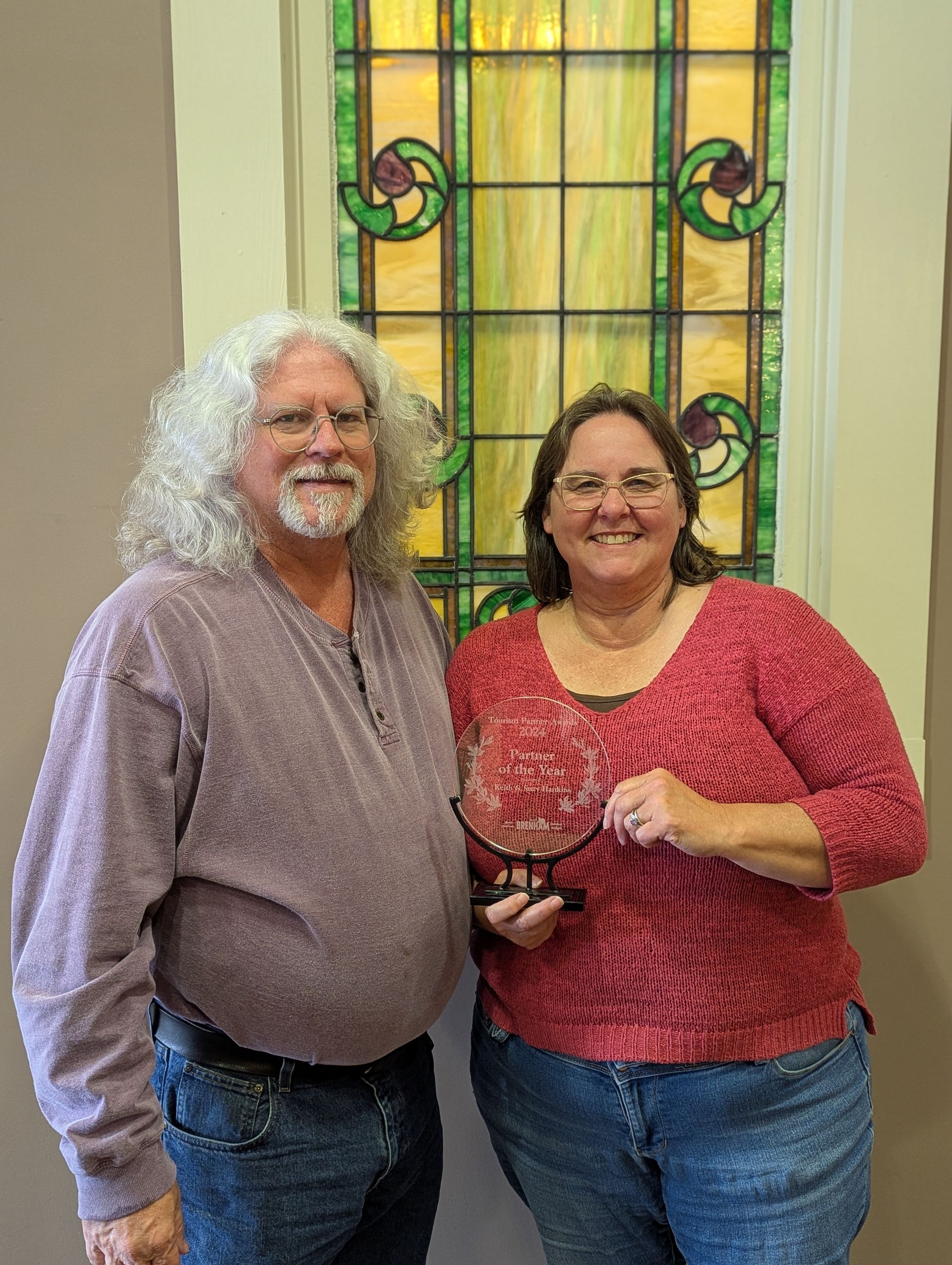 Man and woman smiling, holding a trophy, in front of stained glass window.