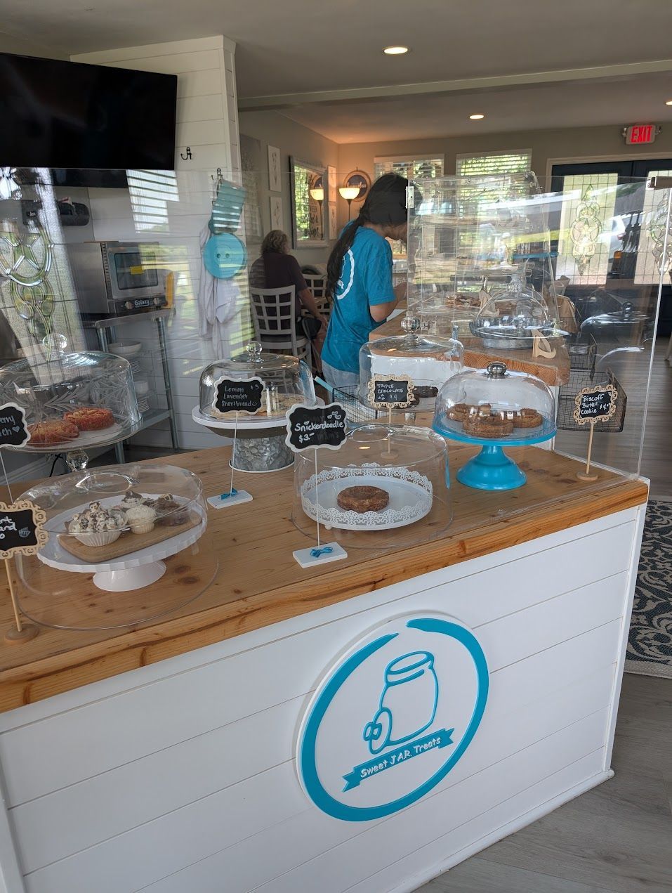 A bakery counter displays baked goods. A person in a blue shirt stands behind the counter, serving customers.