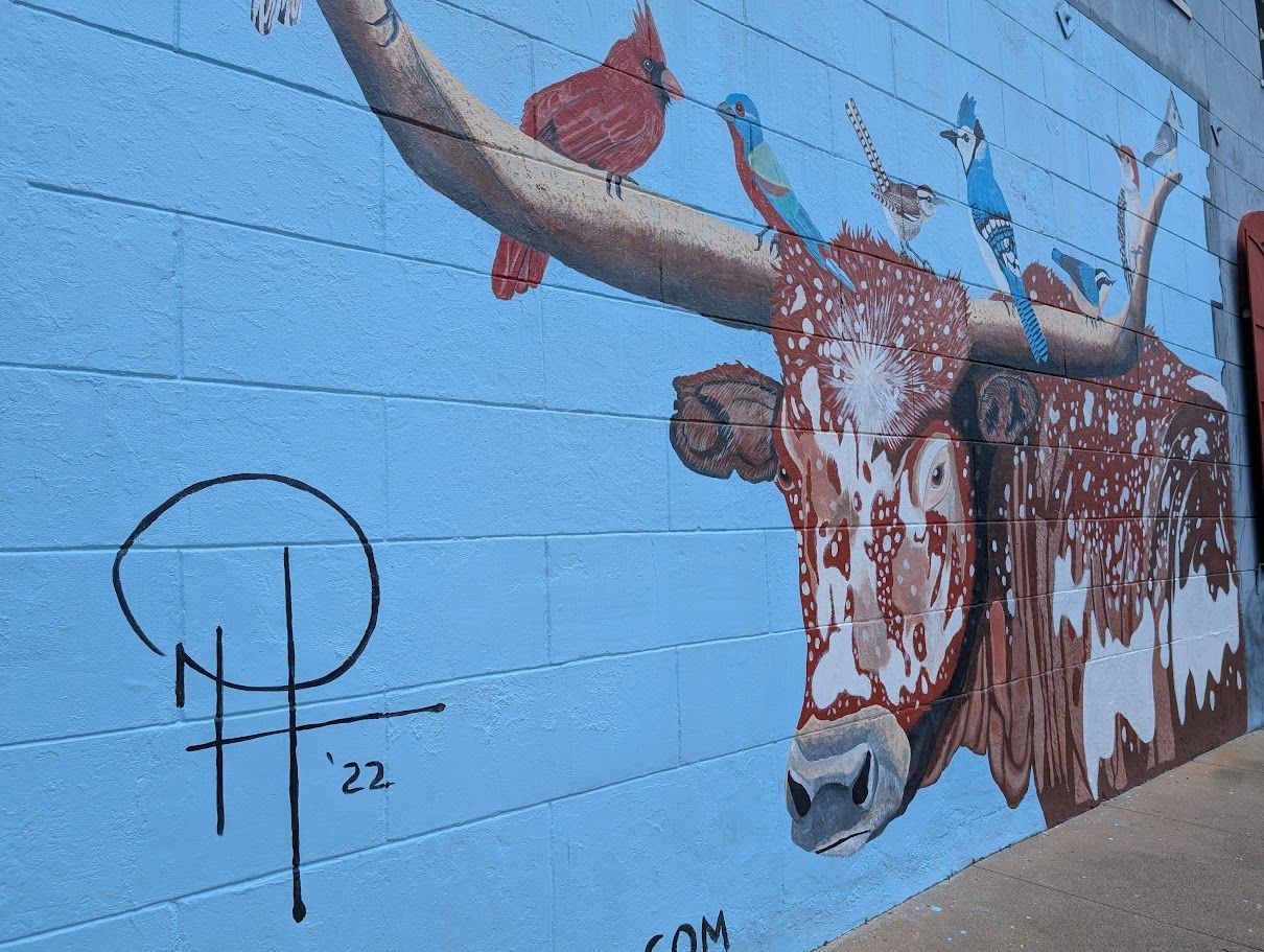 Mural of a longhorn with birds on its horns against a blue sky background. Signed by the artist.
