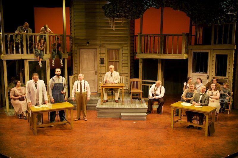 Stage set of a play with people at tables, on balconies, and on a porch. Red and orange backdrops.