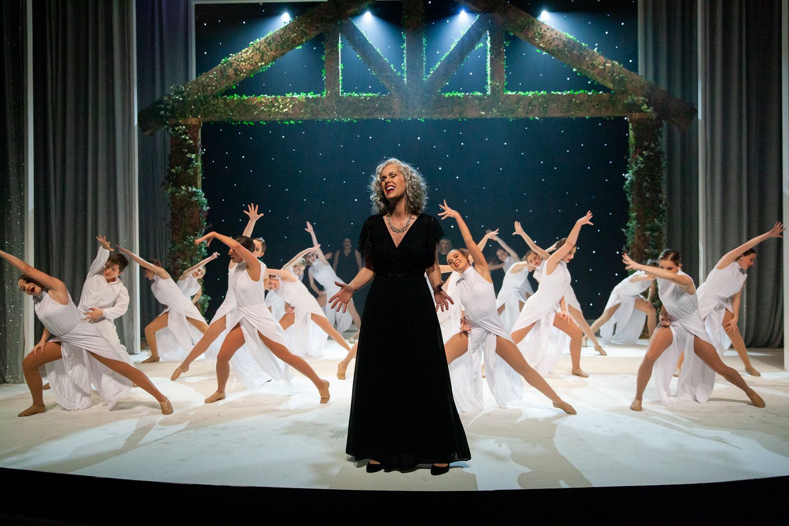 A woman in a black dress is standing in front of a group of dancers on a stage.