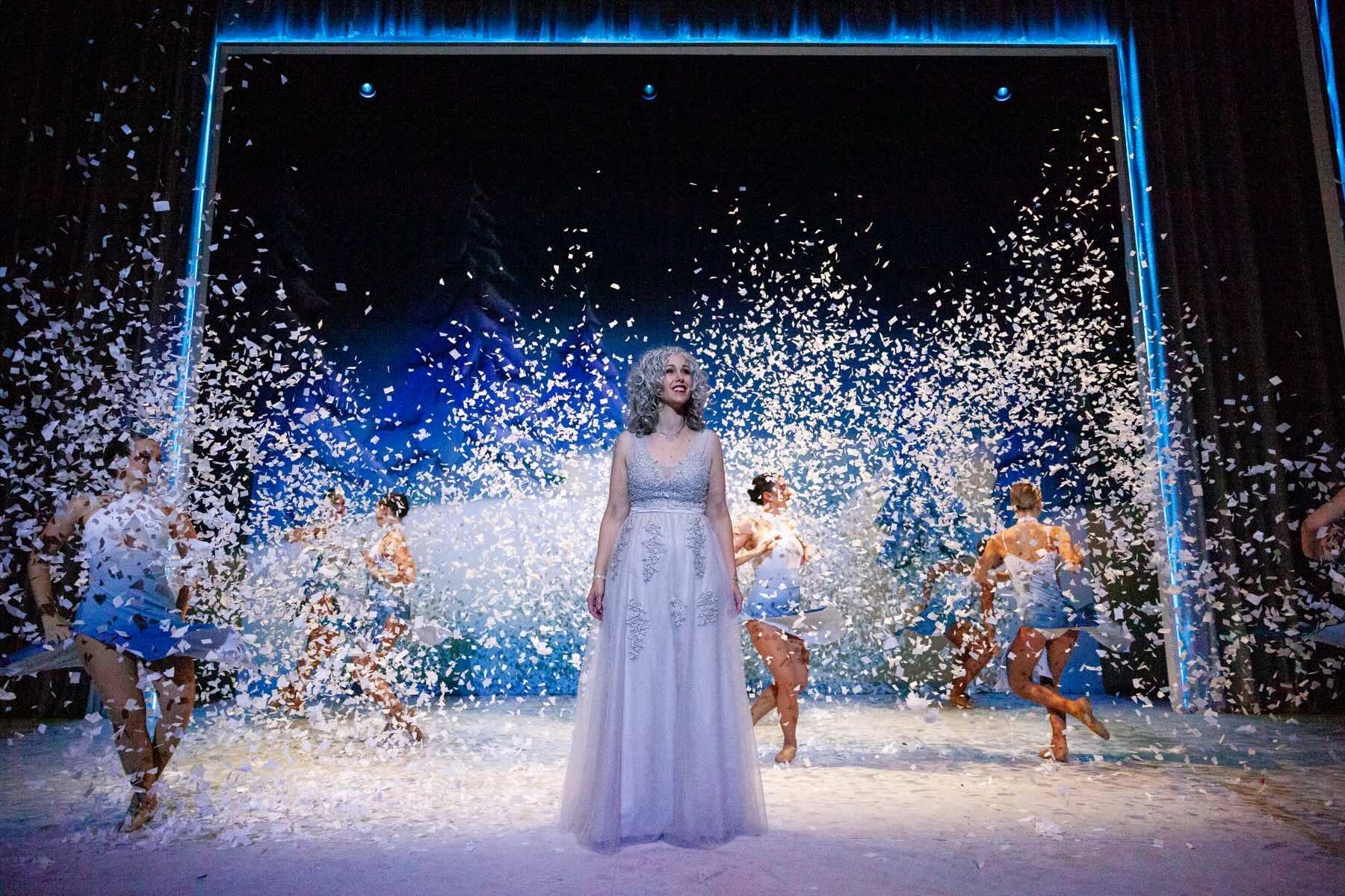 A woman in a white dress is standing on a stage with snow falling around her.