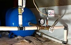 A blue tank is sitting next to a gauge and pipes in a basement.