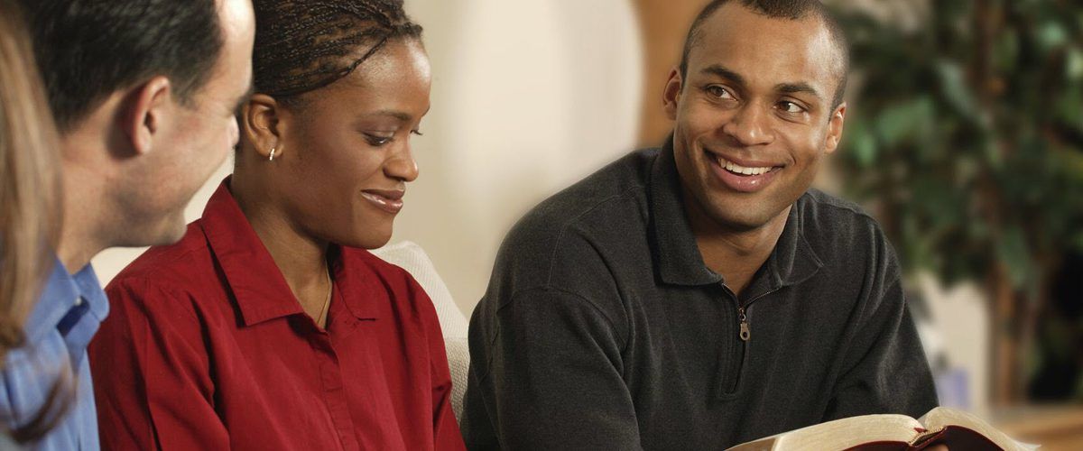 People seated around a white table, holding books and talking. Room has round tables and red chairs.