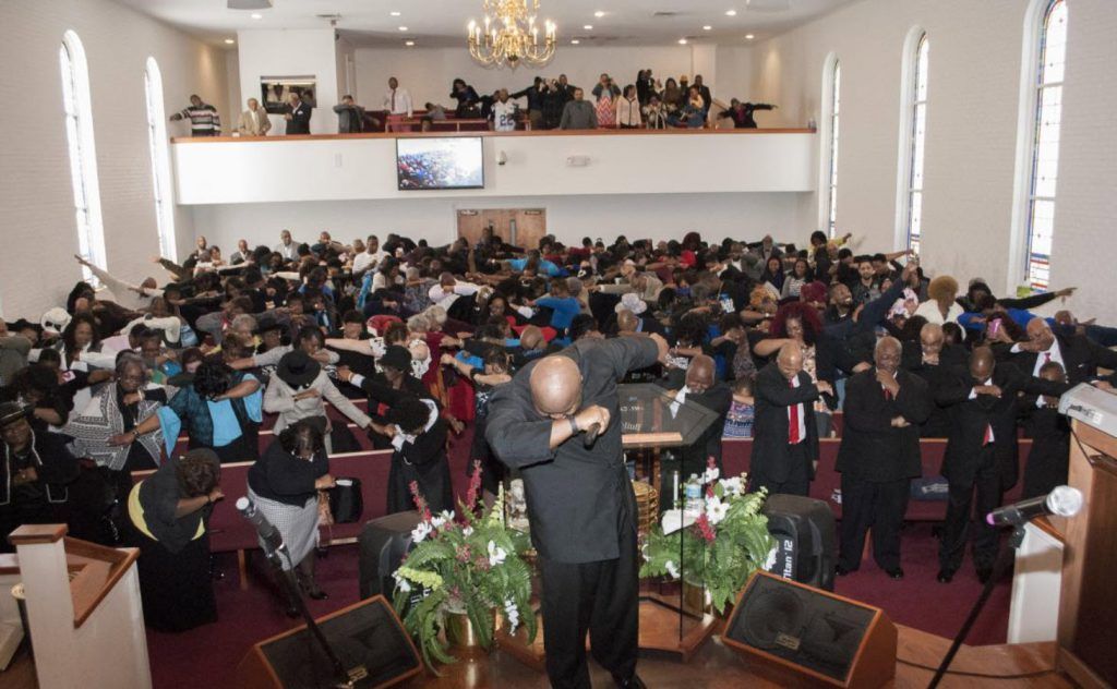 People seated around a white table, holding books and talking. Room has round tables and red chairs.