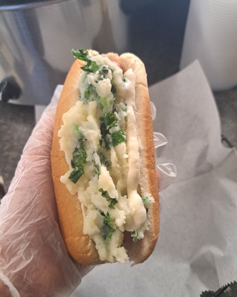 A person wearing gloves is holding a hot dog with mashed potatoes and greens
