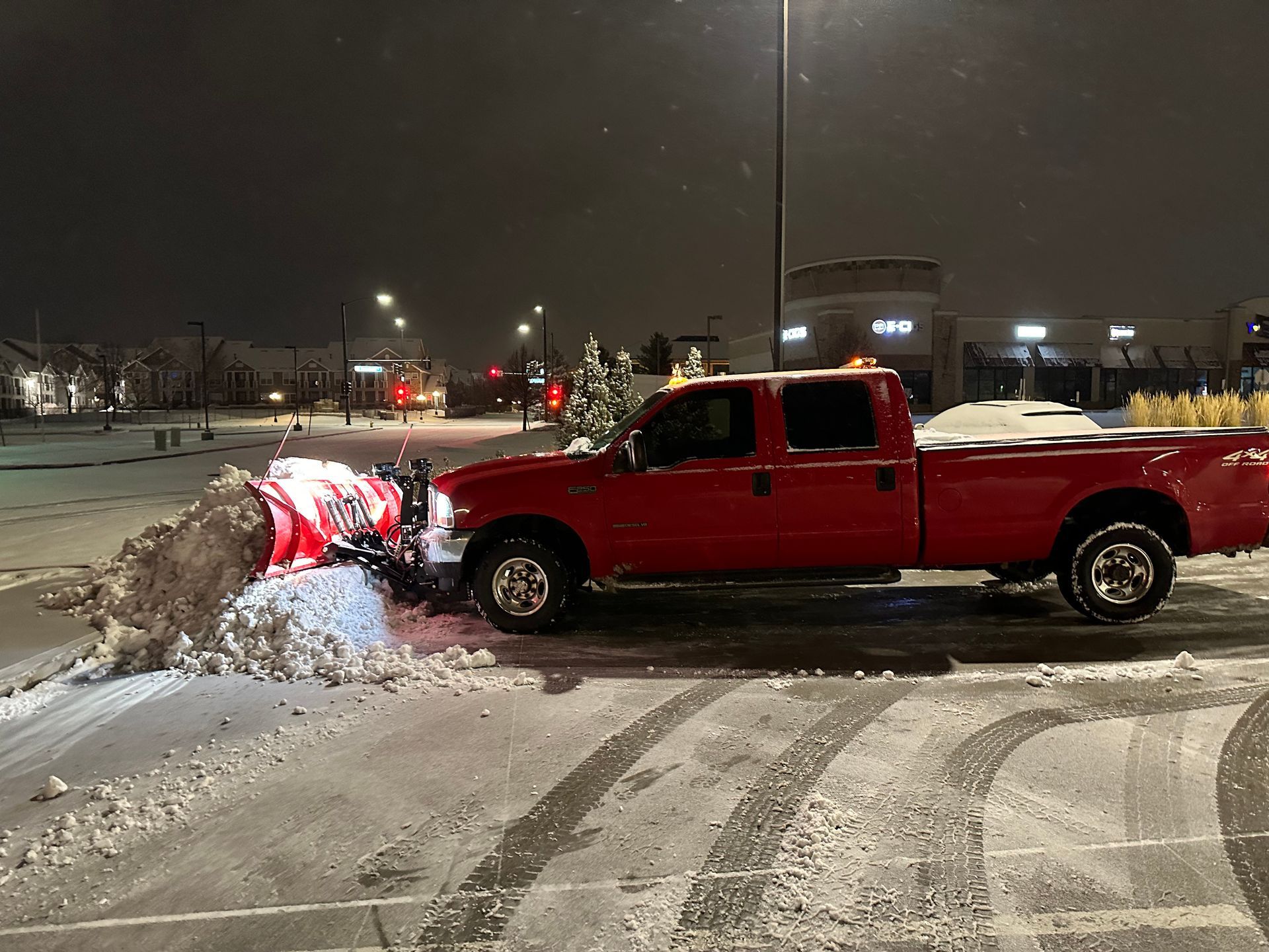 A red truck is plowing snow in a parking lot at night.