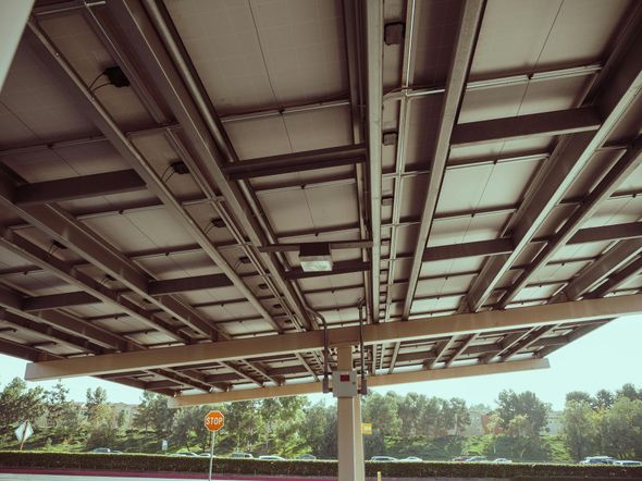 Underside view of solar panels installed over a parking area. Metal framework supports rectangular panels, white support poles.