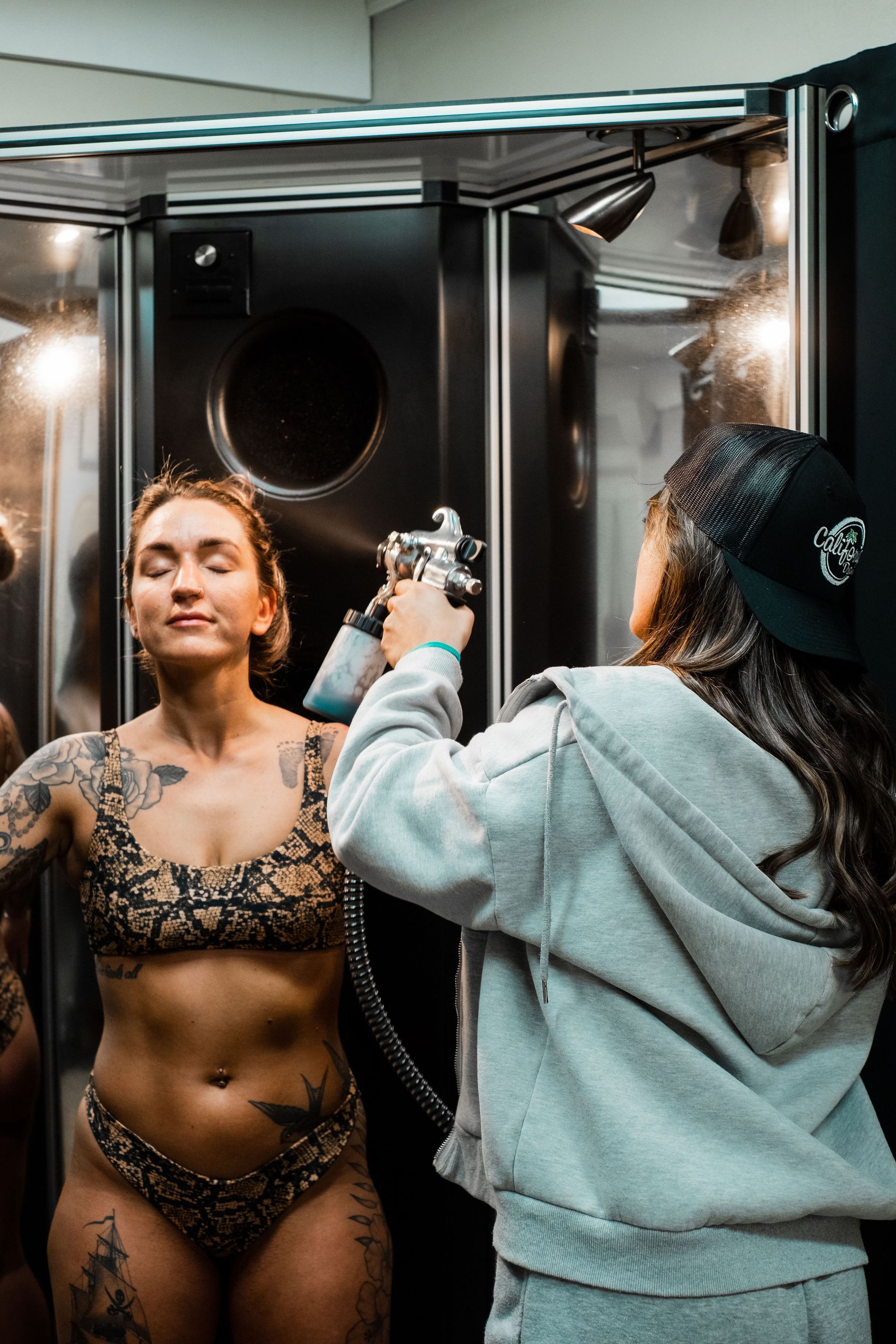 Woman getting spray tan in a booth. Another person sprays her with a tanning gun.