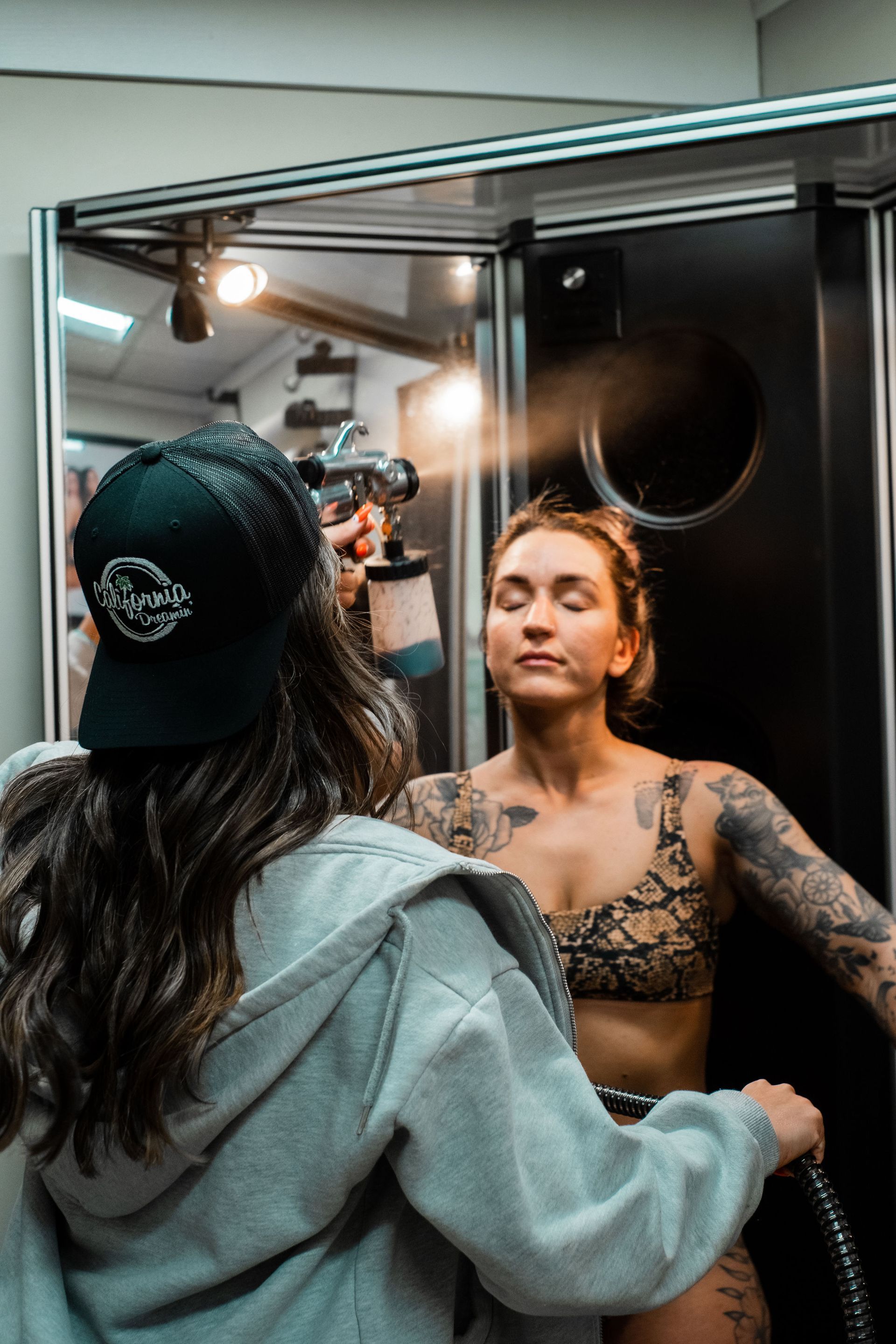 Woman getting spray tan, eyes closed. Person in cap sprays her with a machine in a booth.