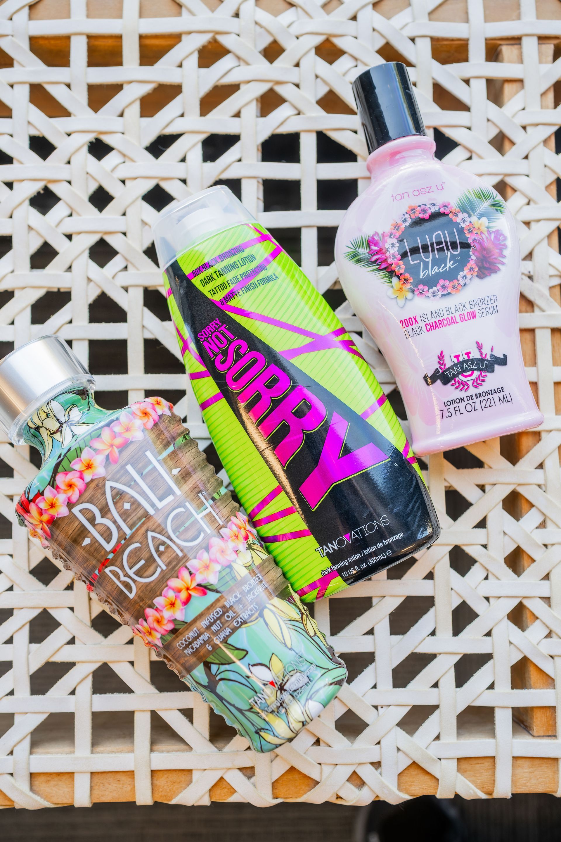 Three bottles of tanning lotion on a woven white surface. The bottles are different colors with floral designs and text labels.