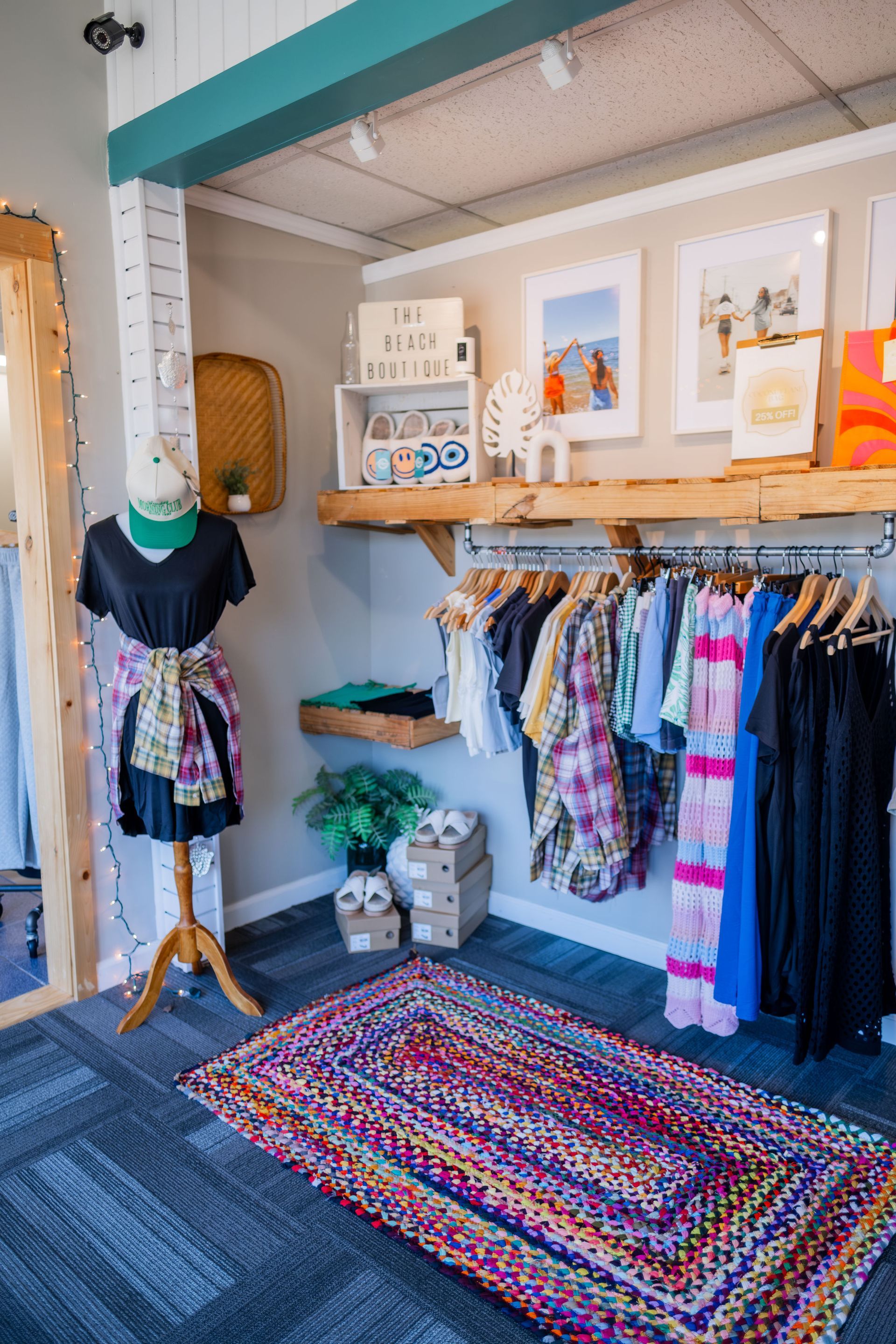 Clothing boutique interior with racks of clothes, a dress on a mannequin, and a colorful rug. Light wood, teal, and white decor.