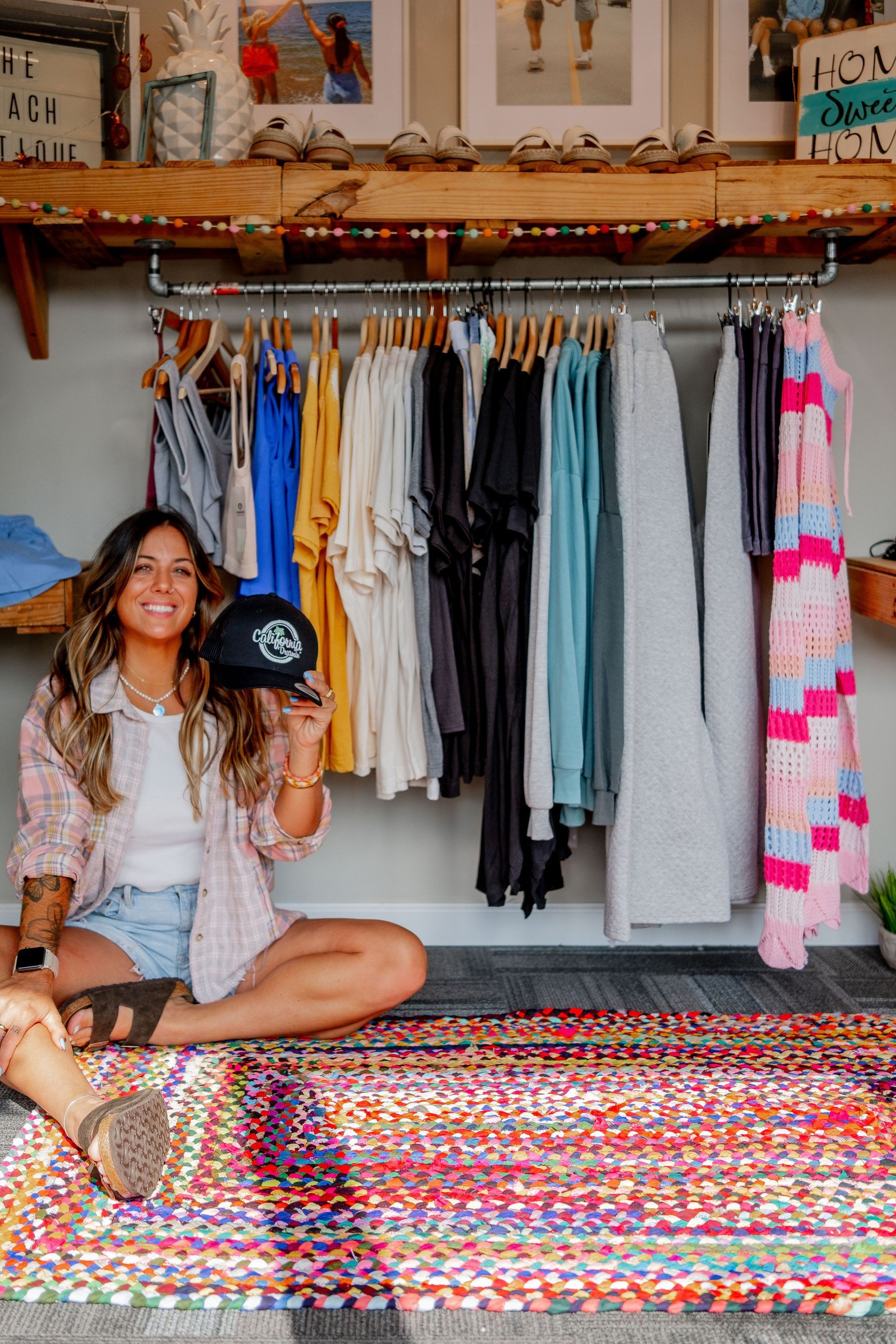 Woman sitting on a colorful rug holding a black hat, with a clothing rack of various colored clothes behind her.