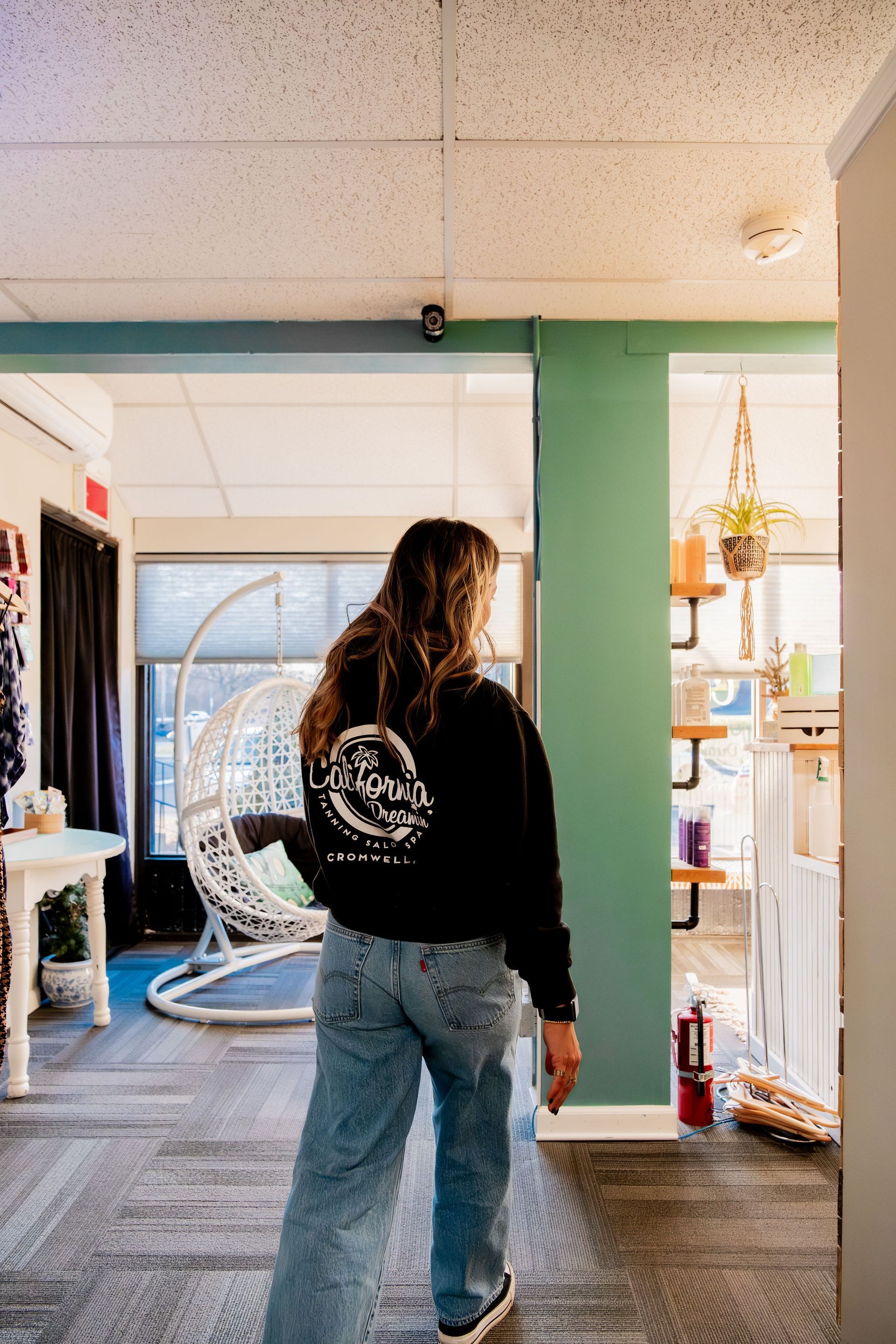 A person in a black sweatshirt and jeans walks down a hallway, looking toward a shop area with plants and shelves.