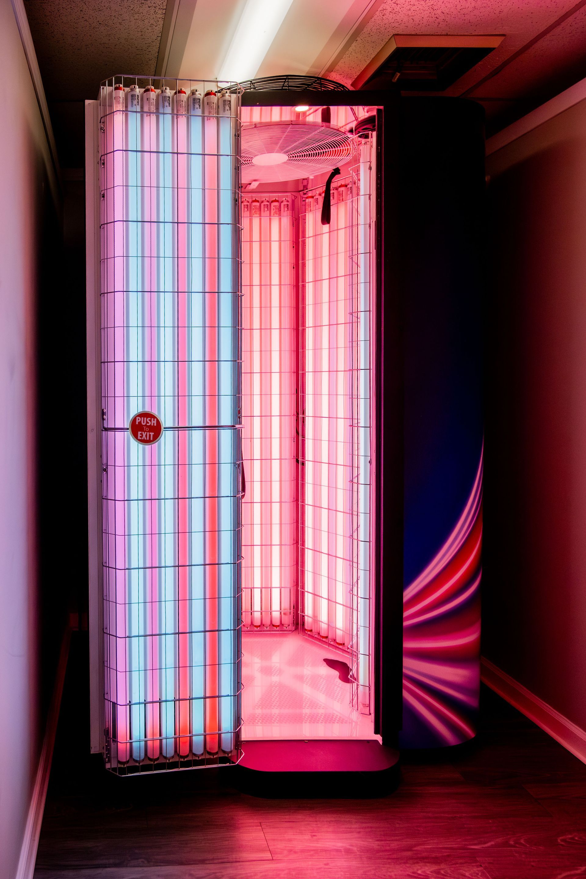 Tanning booth with pink and red UV lights illuminating the interior. The door is open, set in a narrow hallway.