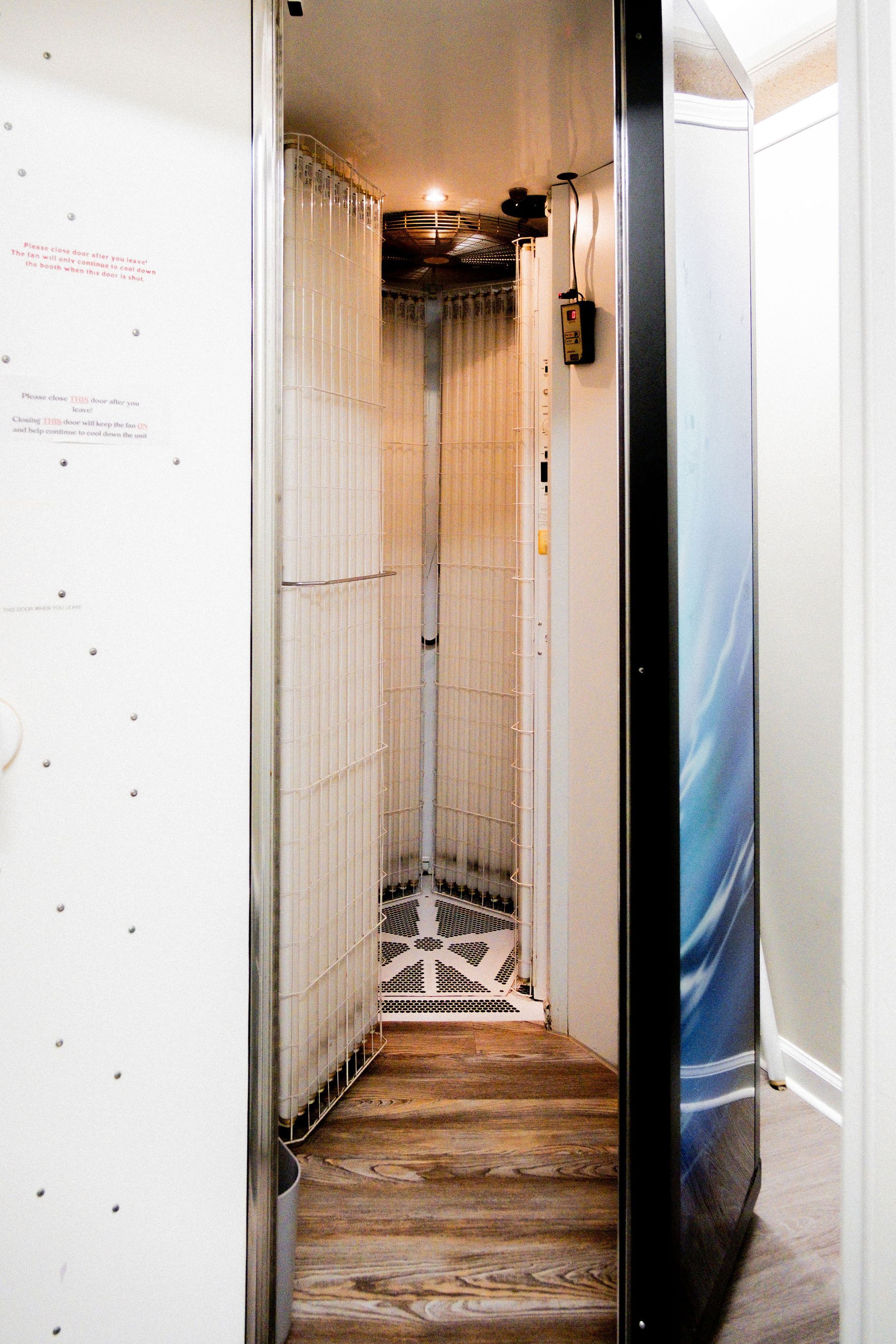 Narrow elevator interior with wooden floor and metal framework. White walls, exposed mechanics at ceiling.