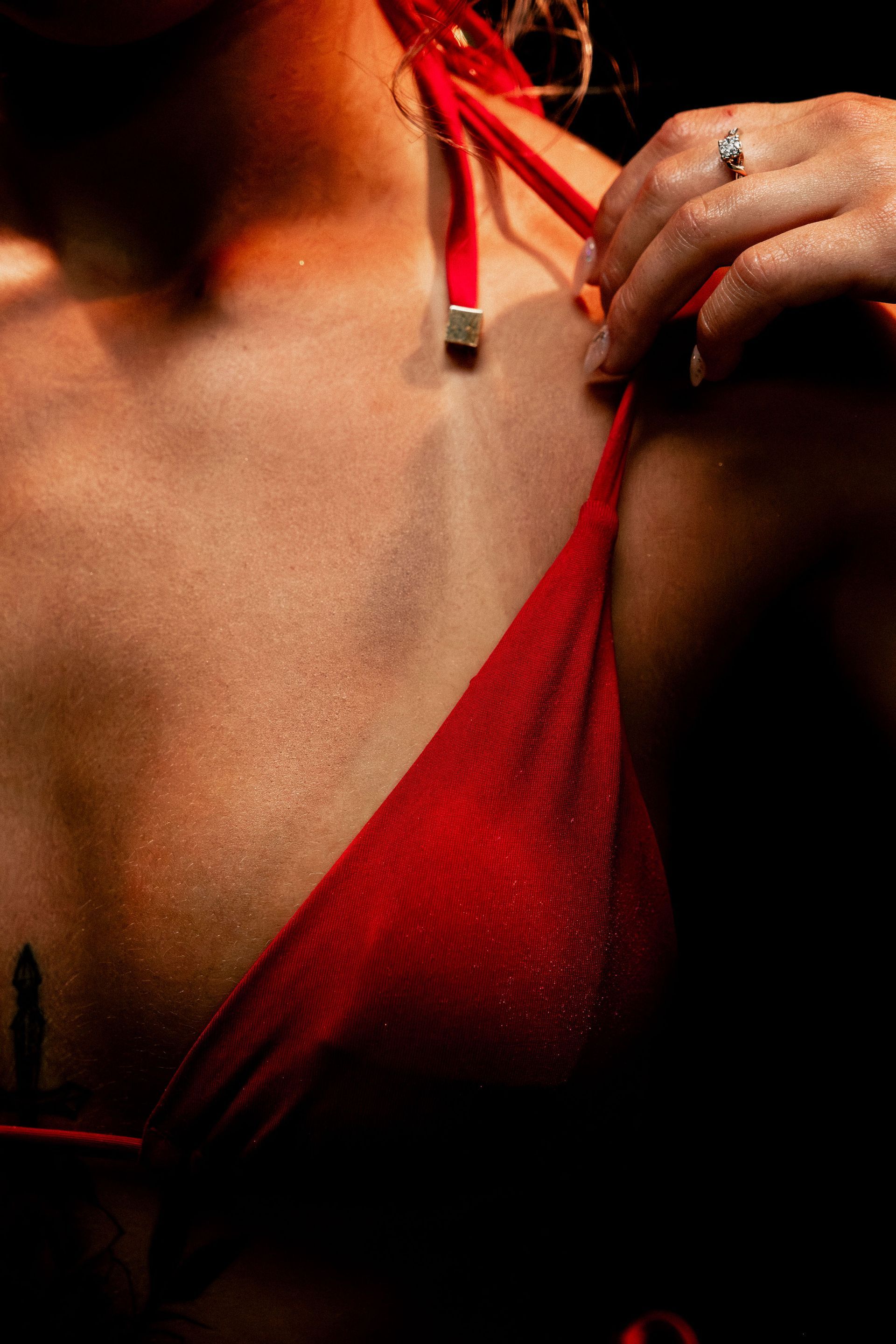 Woman in red bikini top, adjusting strap. Hand with ring visible, skin highlighted, dark background.