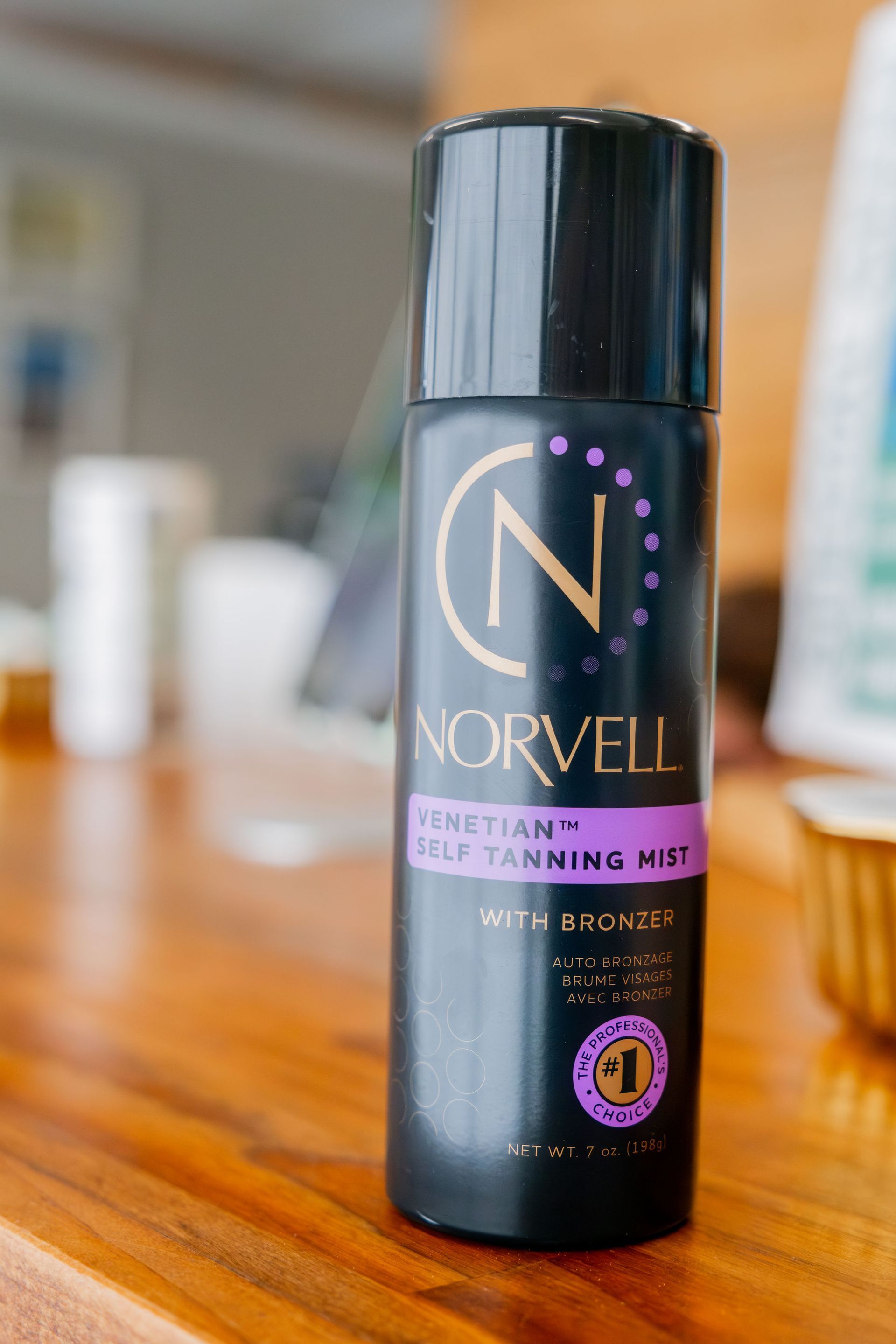 A Norvell Venetian self-tanning mist bottle on a wooden table. It's black with gold and purple details and a gold 