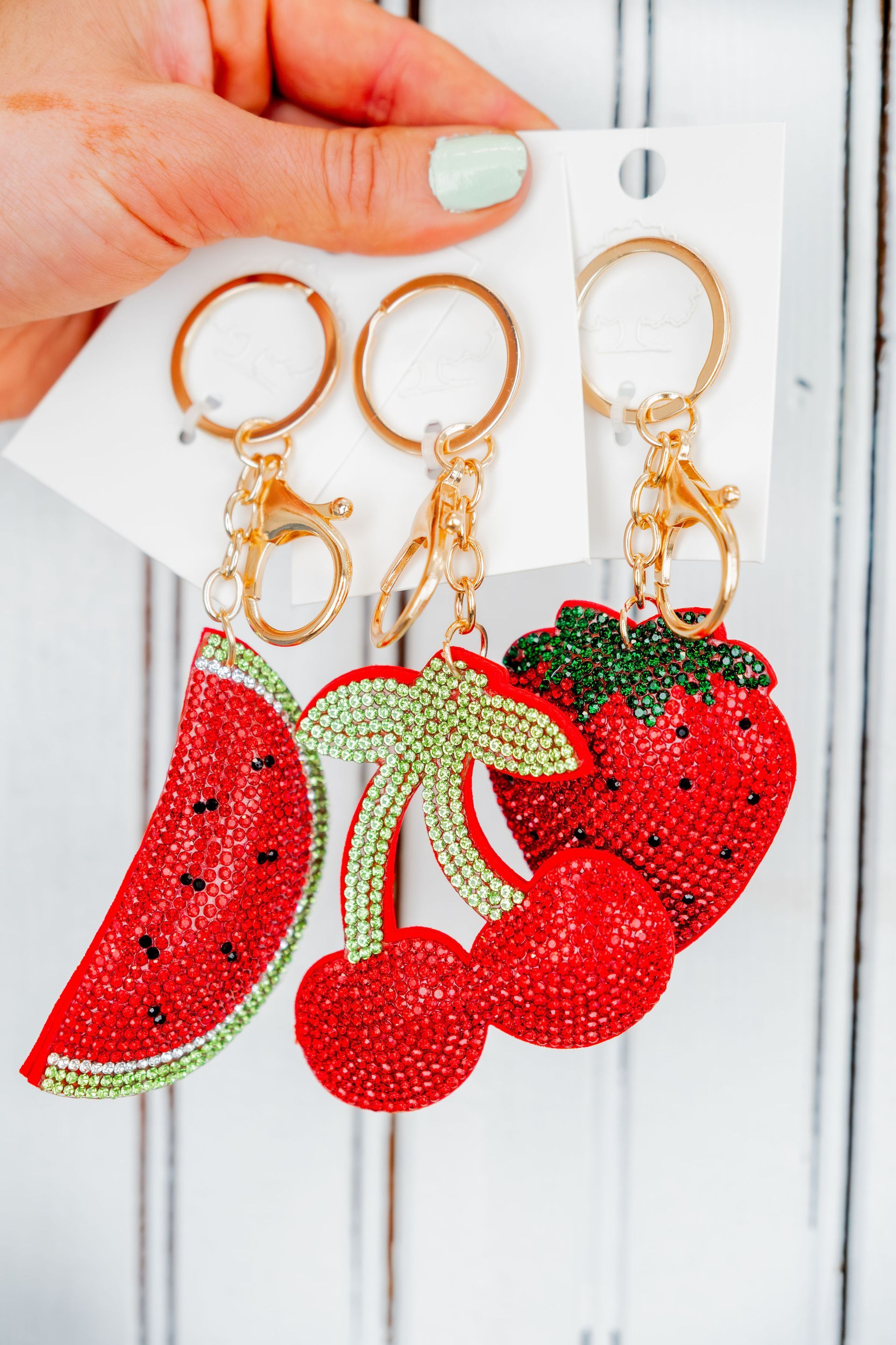A hand holding three keychains with bejeweled fruit charms: a watermelon slice, cherries, and a strawberry.