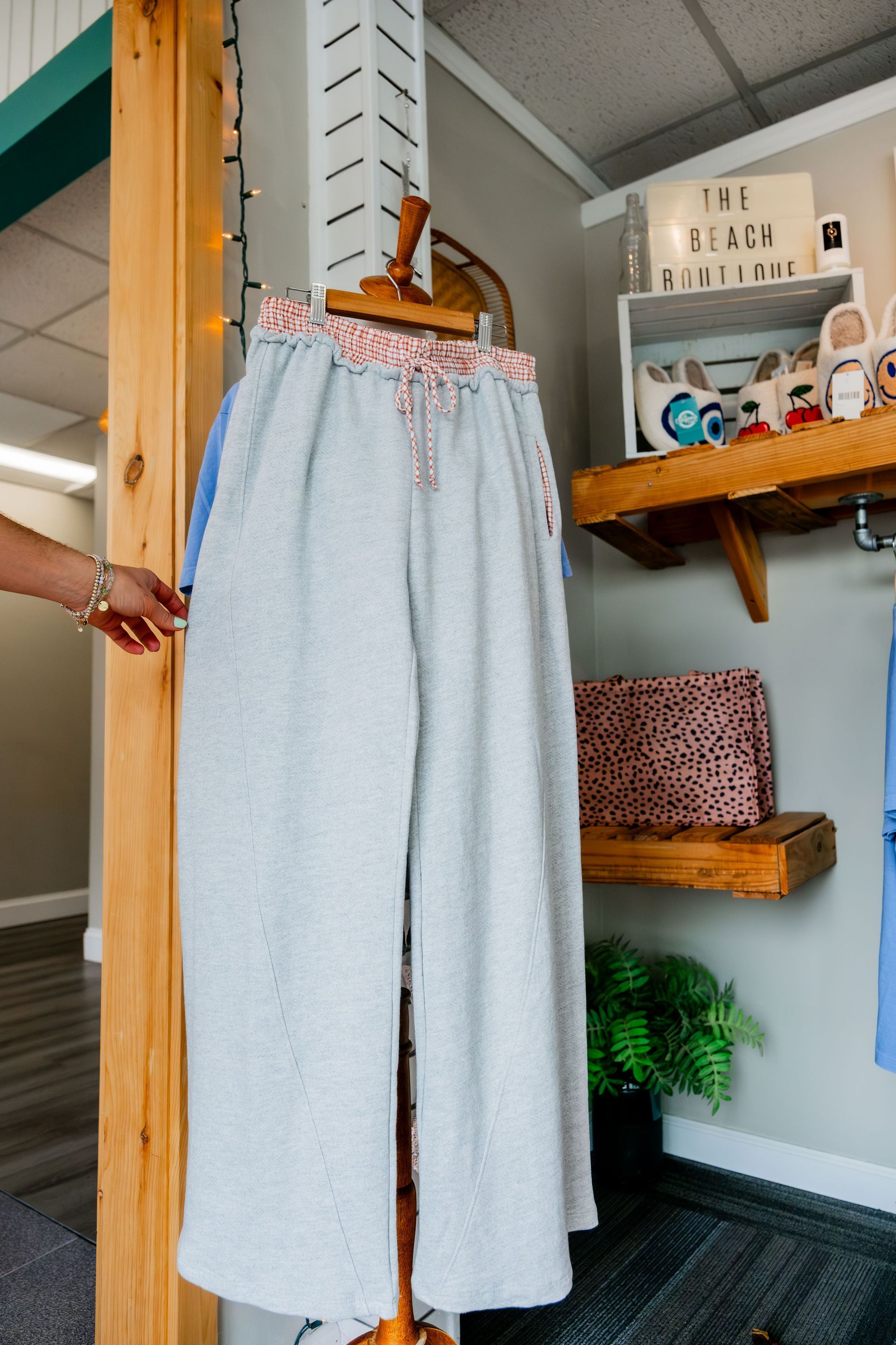 Gray wide-leg sweatpants hanging on a wooden rack inside a retail store. A person's hand touches the pants.