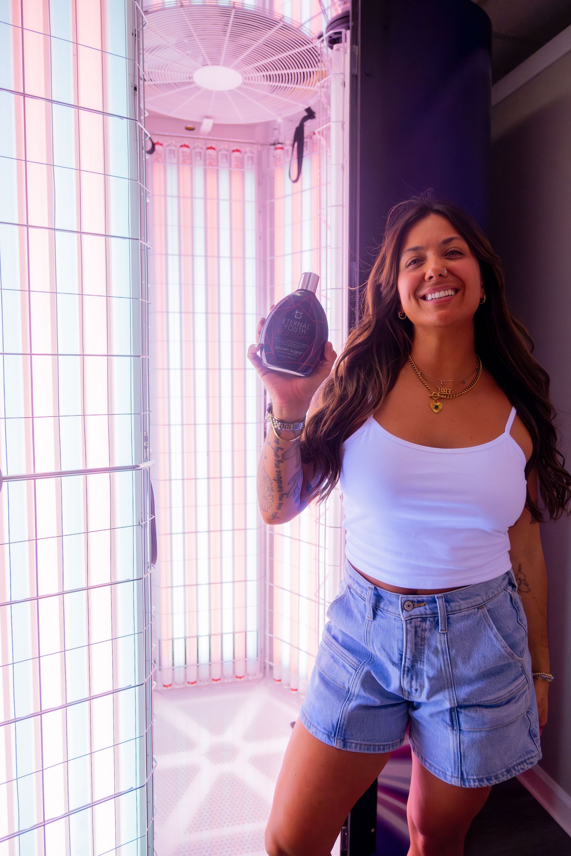 Woman smiles, holding a black spray bottle near a tanning bed; wearing a white tank top and denim shorts.