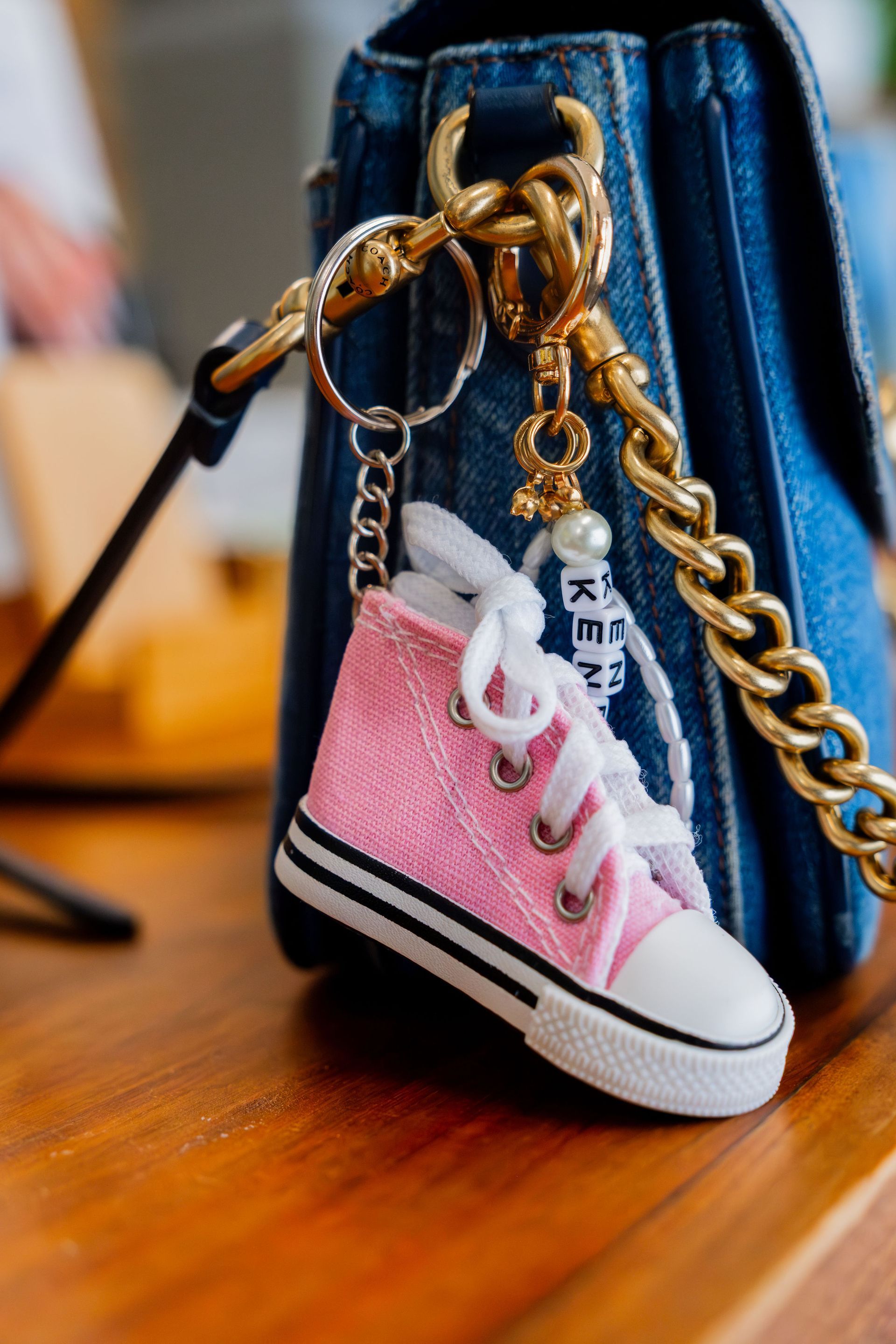 A pink sequined sneaker charm with white laces hangs from a gold chain attached to a denim purse.