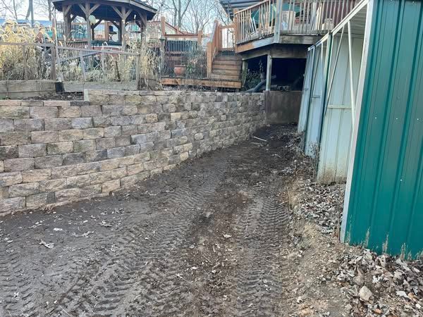 A dirt road leading to a house with a stone wall and a green shed.