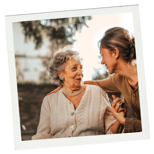 Elderly woman with her arm around another woman in an outdoor setting, both smiling.