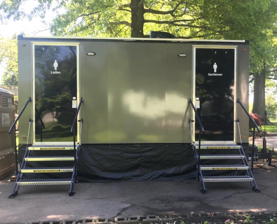 Two portable toilets, side by side on top of the grass in a park with palm trees and a nearby forest