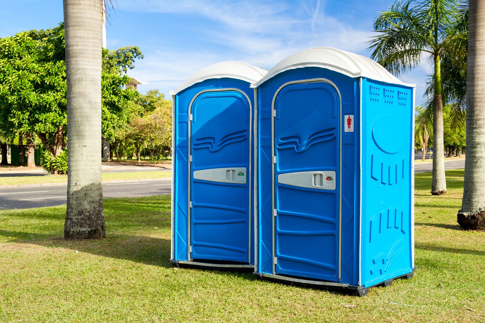 Two portable toilets, side by side on top of the grass in a park with palm trees and a nearby forest