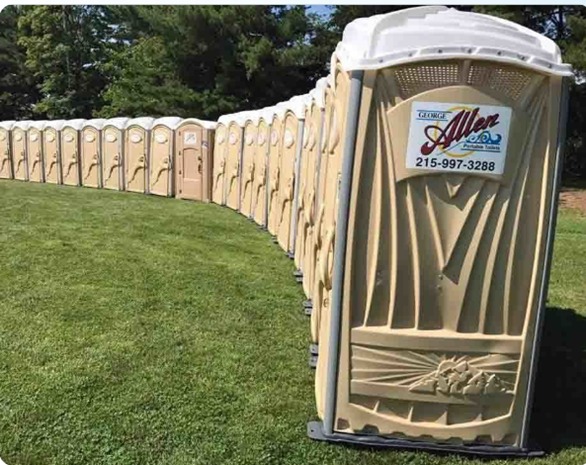 Long row of beige porta-potties lined up neatly on grassy park field.