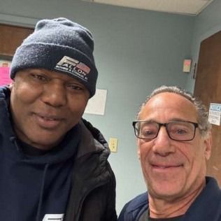 Two men smiling, one in a beanie and jacket, the other with glasses and dark shirt, indoors.