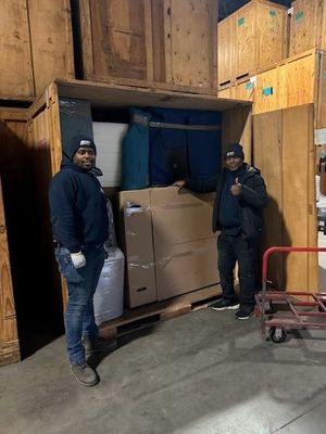 Two movers in a storage unit with packed boxes. One gives a thumbs up.
