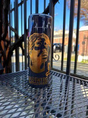 Tumbler with a gold portrait of a person on a black background, outdoors on a metal surface.