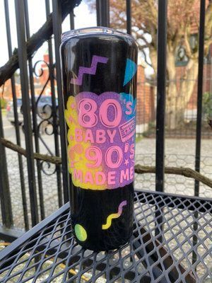 Black tumbler with colorful '80s/'90s design: text, shapes. Outdoors, on a metal surface.