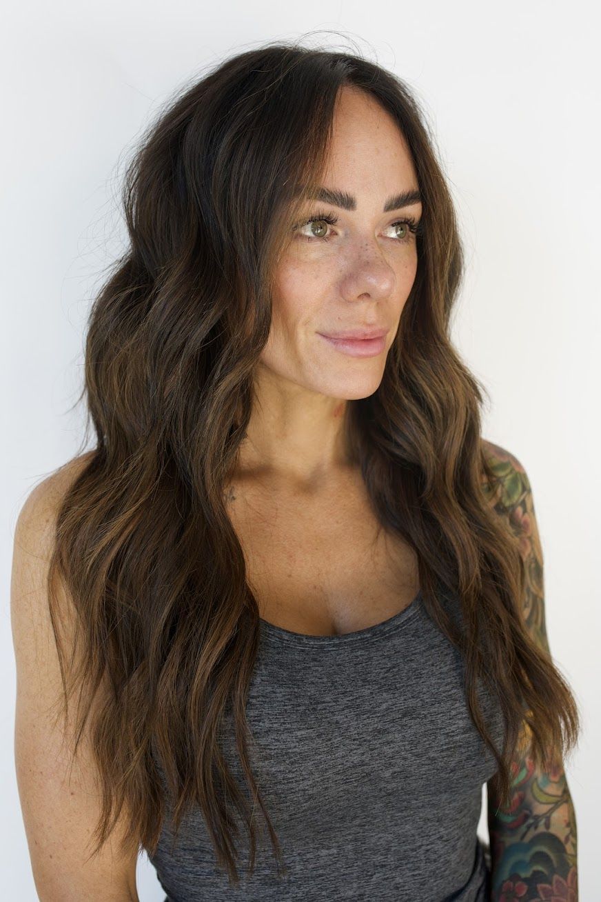 A woman with long brown hair is wearing a grey tank top and has a tattoo on her arm.