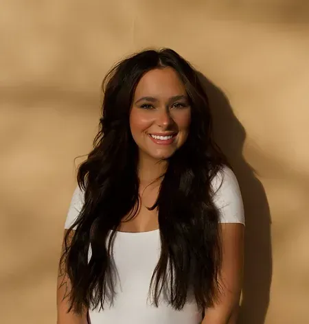 A woman with long dark hair is smiling and wearing a white shirt