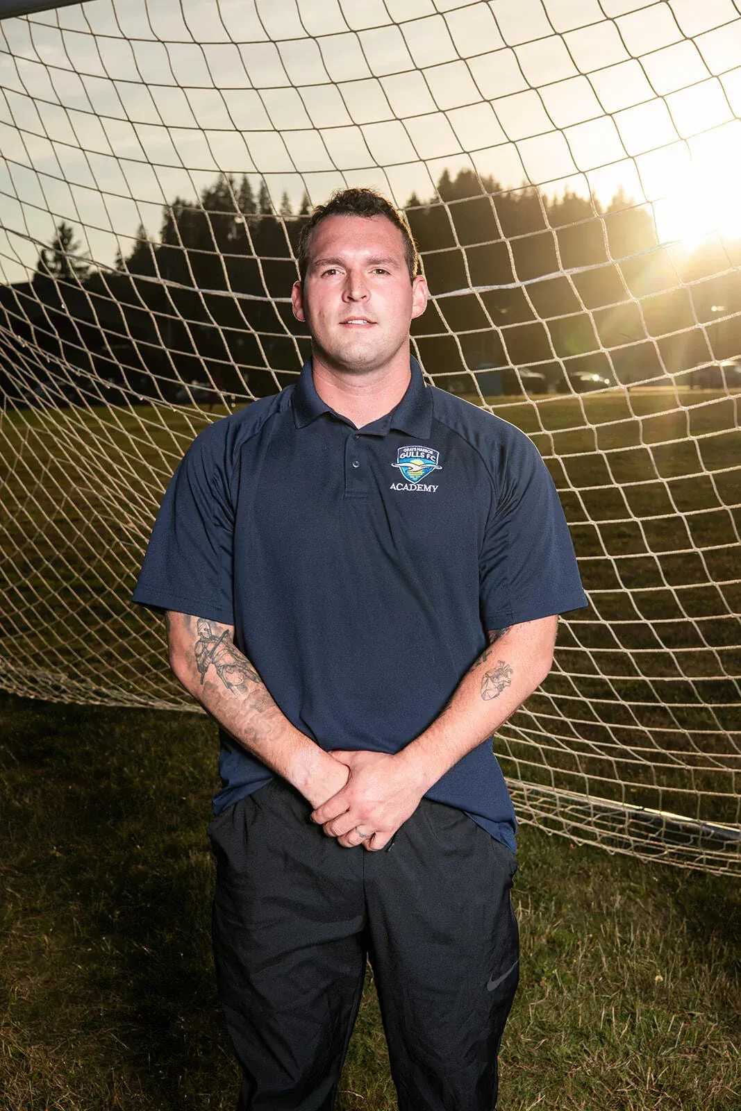Man in navy polo and black pants, standing on a soccer field in front of a net, arms crossed.