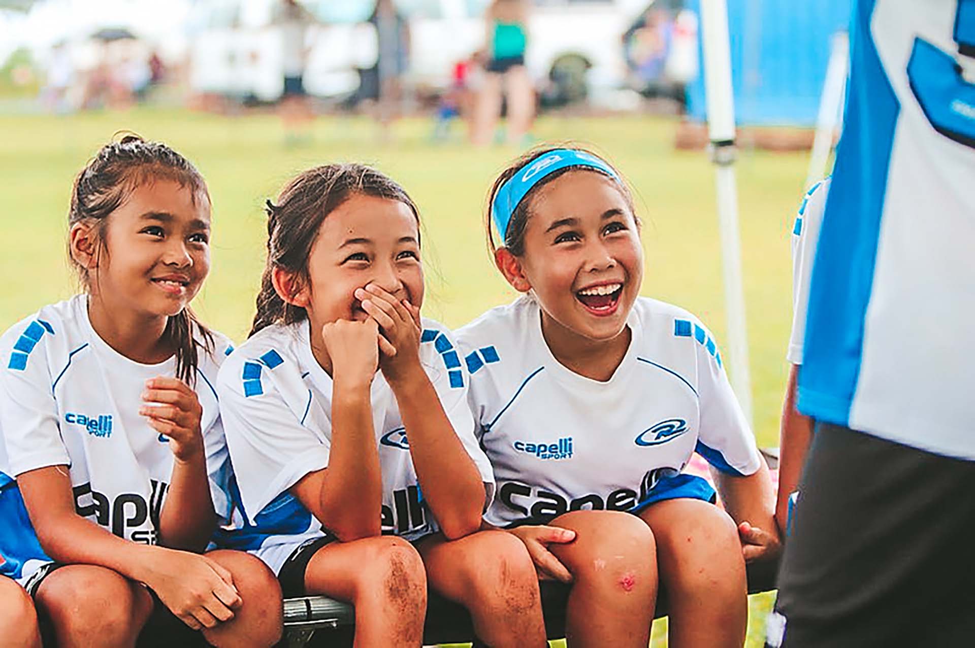 Three soccer players laughing on a bench, wearing white and blue jerseys.