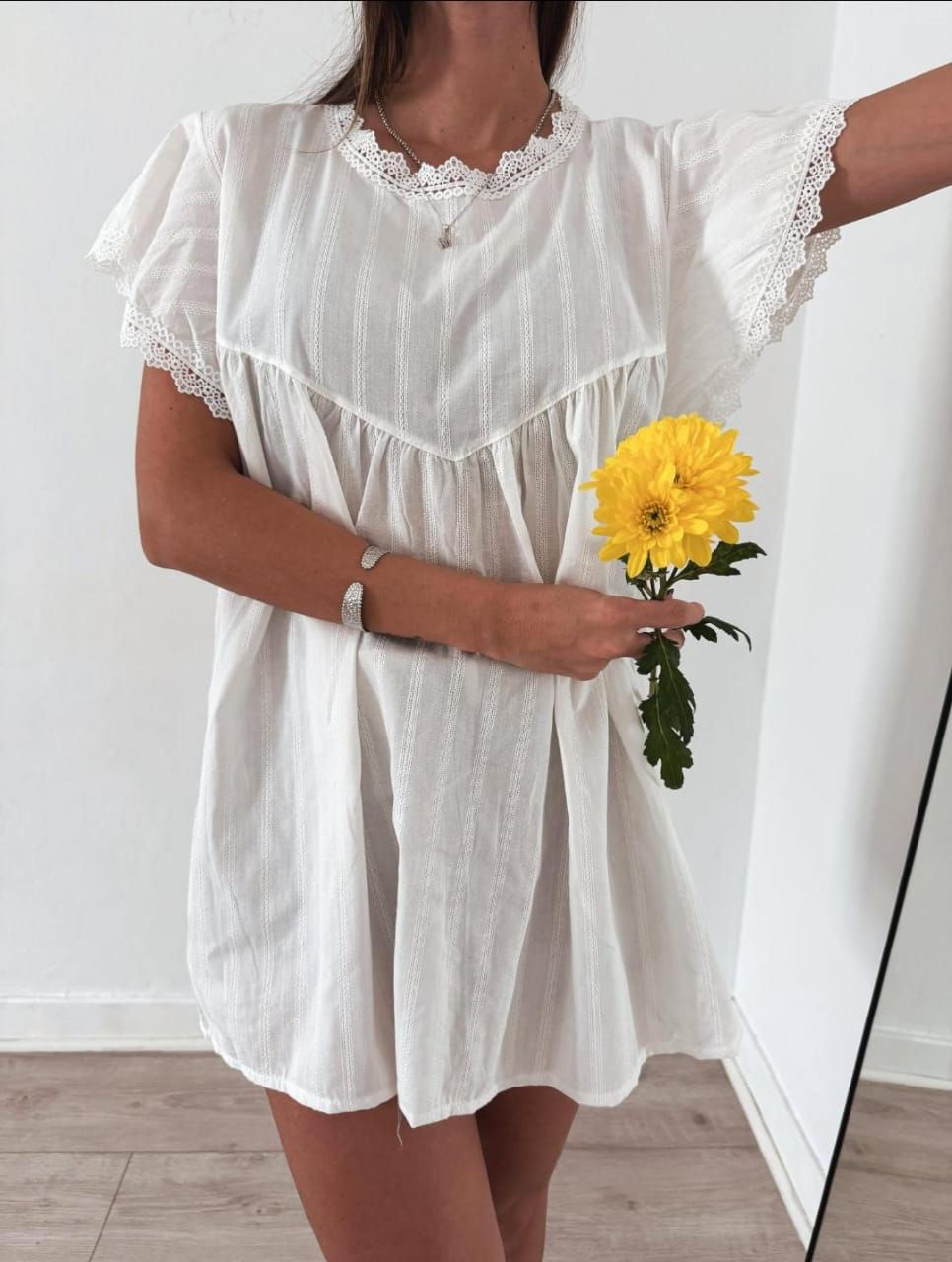 Mujer con vestido blanco sosteniendo una flor amarilla, de pie en la habitación.