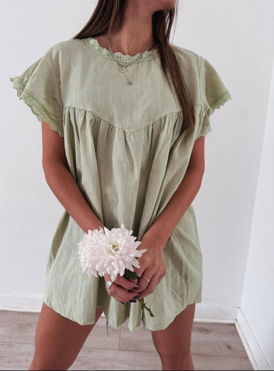 Mujer con un vestido verde claro, sosteniendo una flor blanca. De pie en un interior frente a una pared blanca.